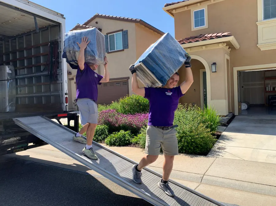 Two Double Move movers carrying wrapped furniture overhead down a truck ramp outside a stucco home